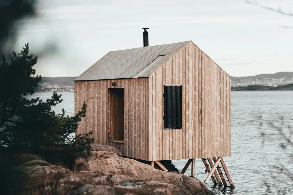 A small pitched-roof sauna clad in pale vertical boards, raised on thin legs over a granite shore, with a ladder down to the water.
