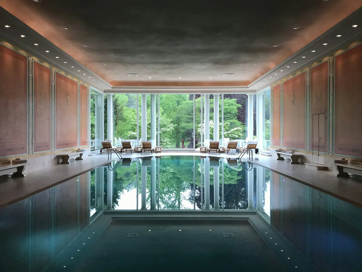 A long indoor pool with still, dark water reflecting a glazed end wall and the trees outside, with loungers along both sides.