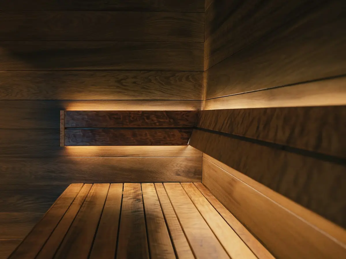 The inside corner of a sauna with warm timber walls, a stepped bench and a strip of light under the upper bench.