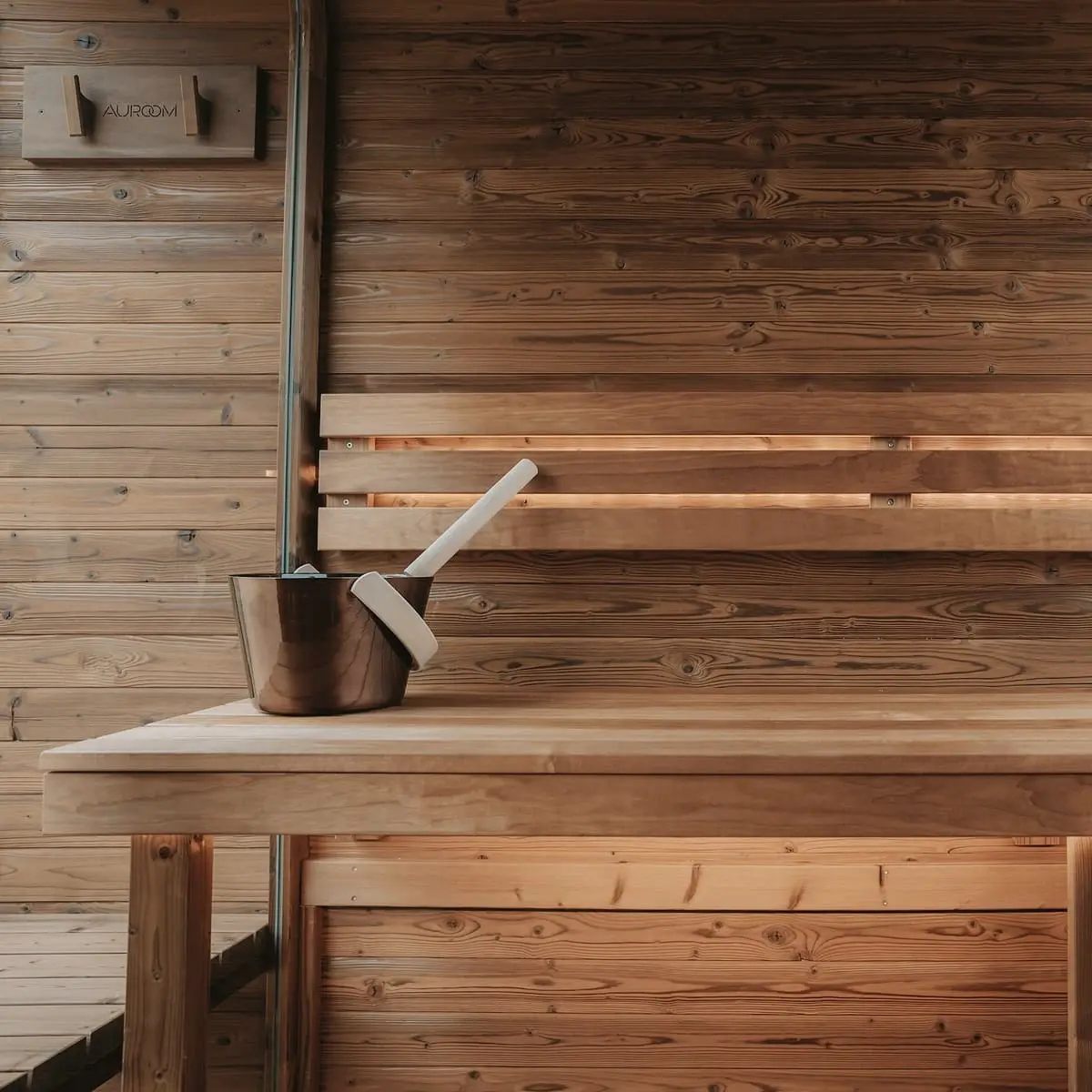 A copper bucket and wooden ladle on a wide timber bench against a pale board wall, with a wall-mounted flue pipe behind.