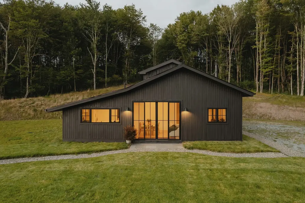 A black timber house with a pitched roof and a deep glazed porch, glowing from inside, on a lawn in front of a wood.