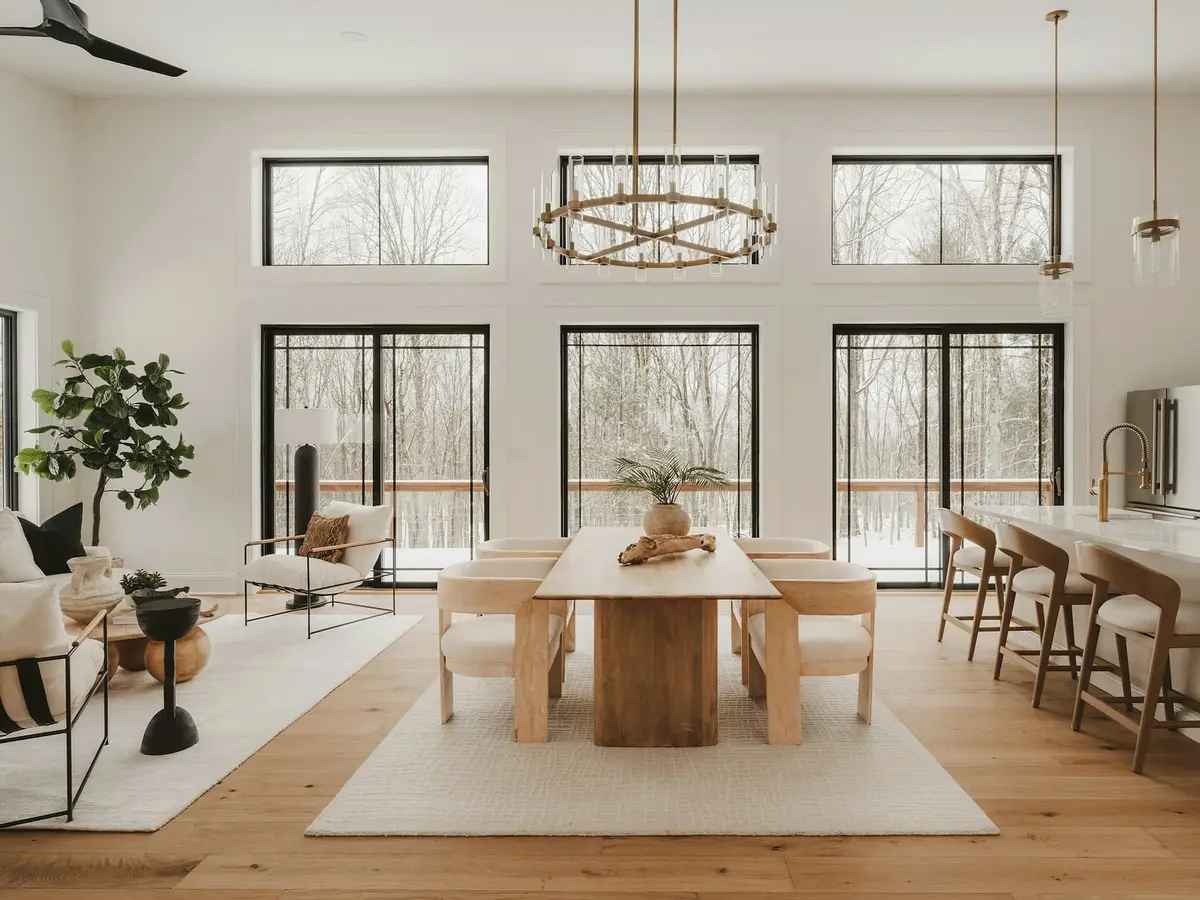 A bright open room with a long oak table, upholstered chairs, a ring chandelier, and three tall black-framed glass doors to a deck.