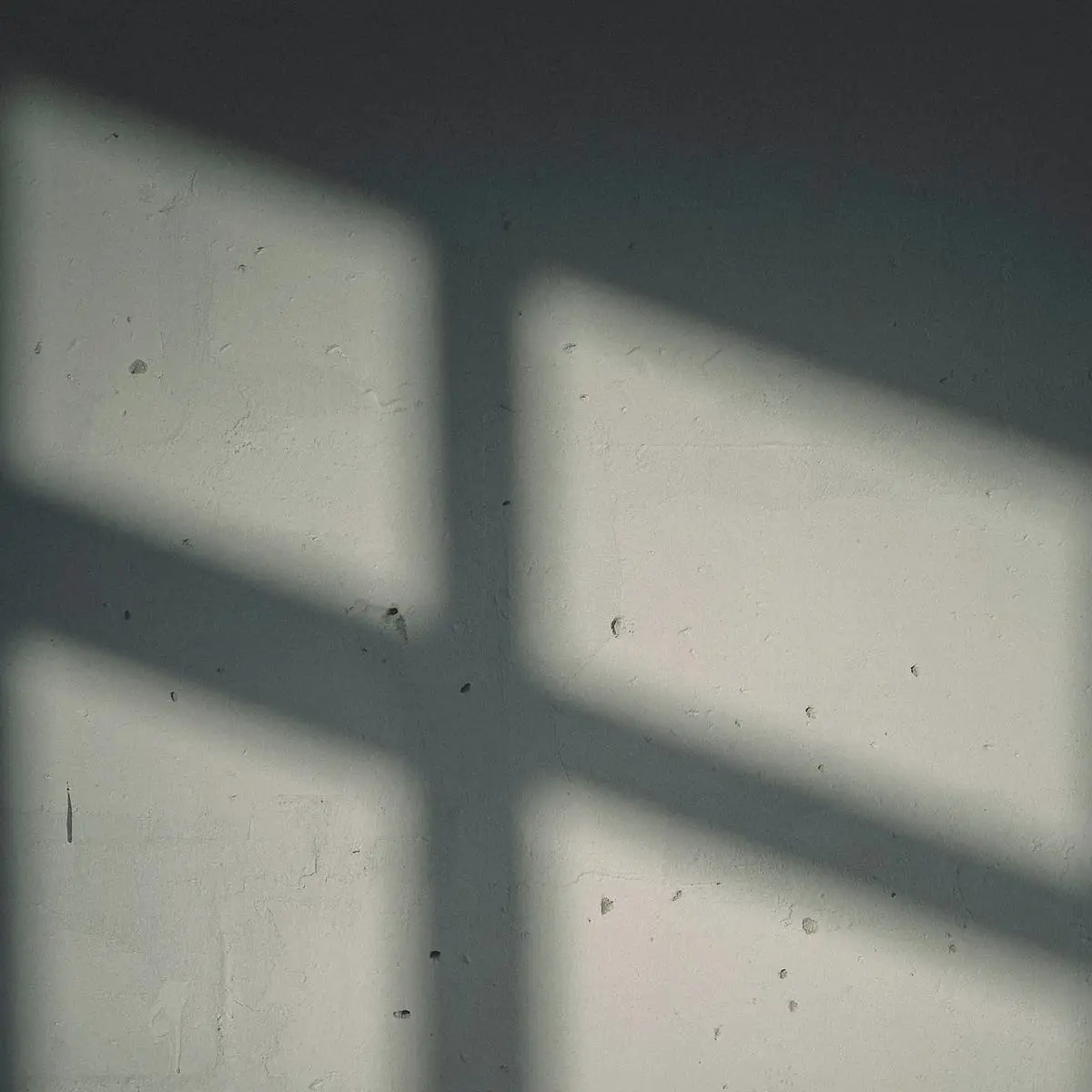 A bare concrete wall with the shadow of a window frame laid across it in grey and cream.