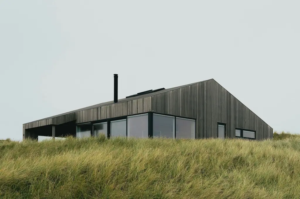 A low house clad in dark timber, with a long pitched roof and a black flue, seen across tall dune grass under a pale sky.