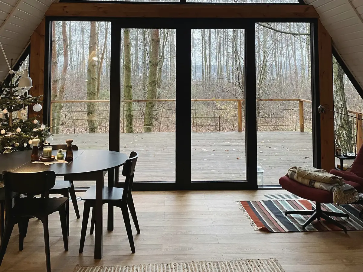 A round dining table with black chairs in front of a tall glazed gable, with bare trees and a timber deck beyond the glass.
