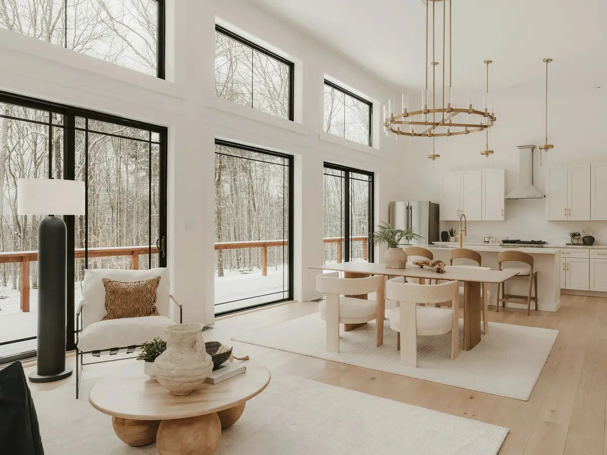 A bright living and dining room with tall black-framed windows looking into a bare winter forest.