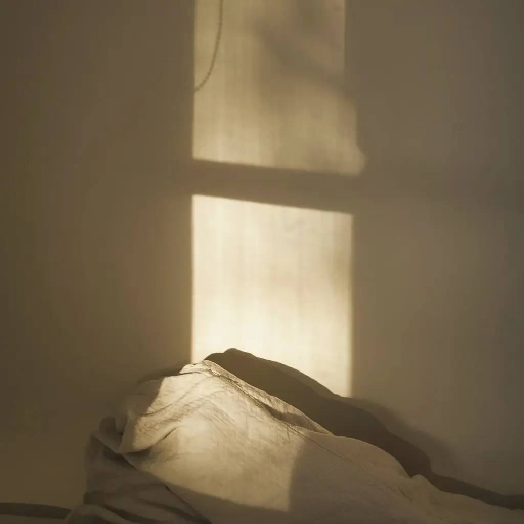 Low sunlight falling through a window onto a pale plastered wall above rumpled linen bedding.