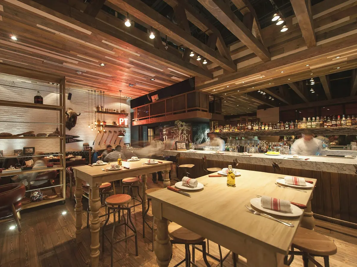 A restaurant with a timber ceiling on beams, a long pale table set for two, a marble-topped open kitchen counter and cooks at work behind it.