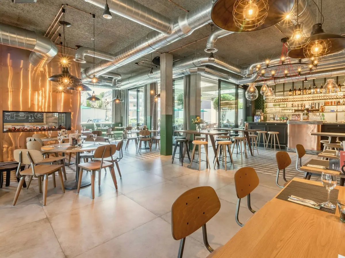 The restaurant dining room with silver ducts, round-backed oak chairs, a long table in the foreground and a lit back bar.