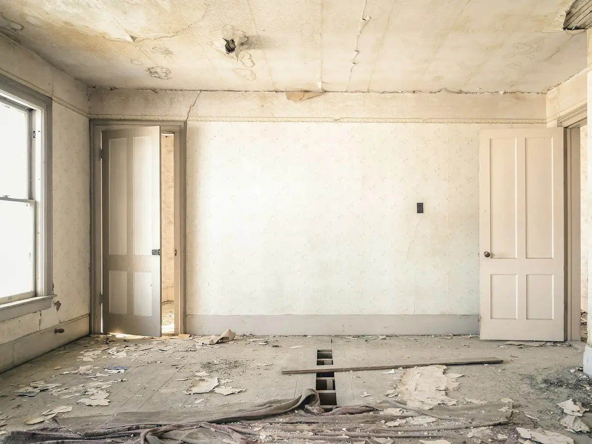 An empty room with cream walls, two tall doors and a sash window, litter and torn flooring across the floor.