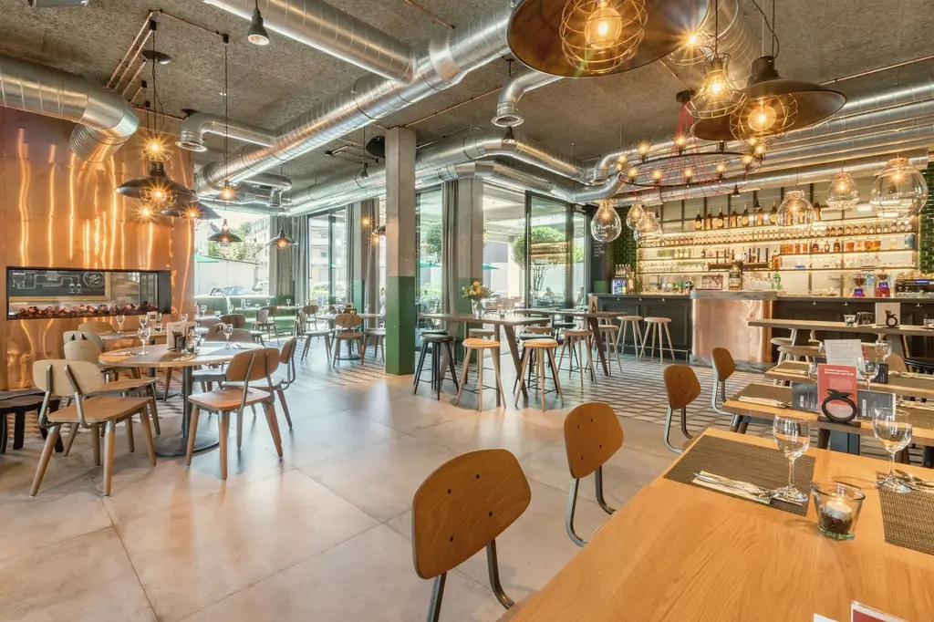 A restaurant with exposed silver ducts under a grey ceiling, a green column, oak chairs with round backs, a long oak table and a lit bar at the back.