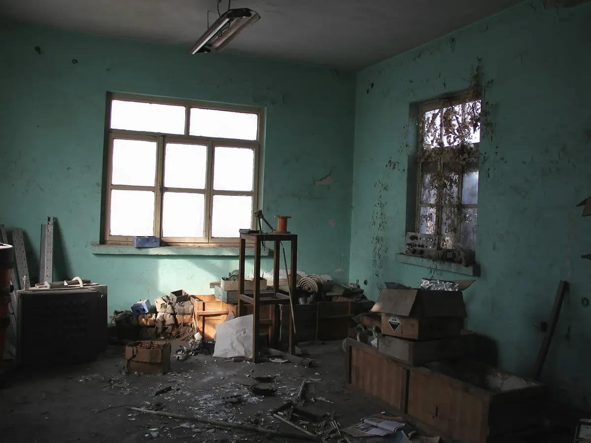 A small room with turquoise painted walls, two large windows, boxes, old shelves and rubbish on the floor.