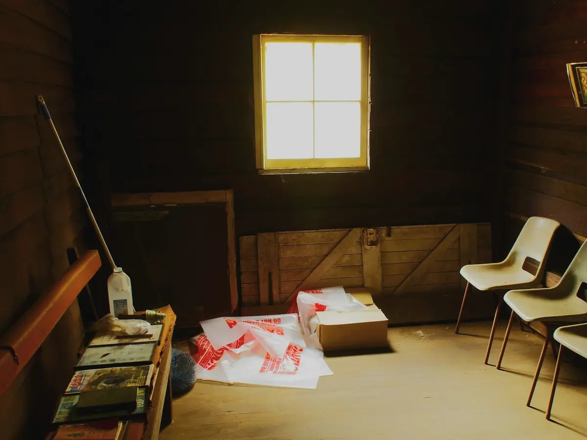 A dark timber-lined room with one small bright window, two plastic chairs, packaging on the floor and a long shelf on the left.