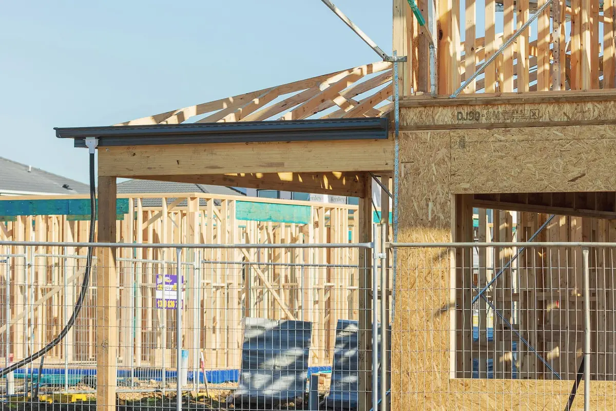 The frame of a house porch and the first lengths of roof truss, with a temporary fence in front and an oriented-strand board wall on the right.