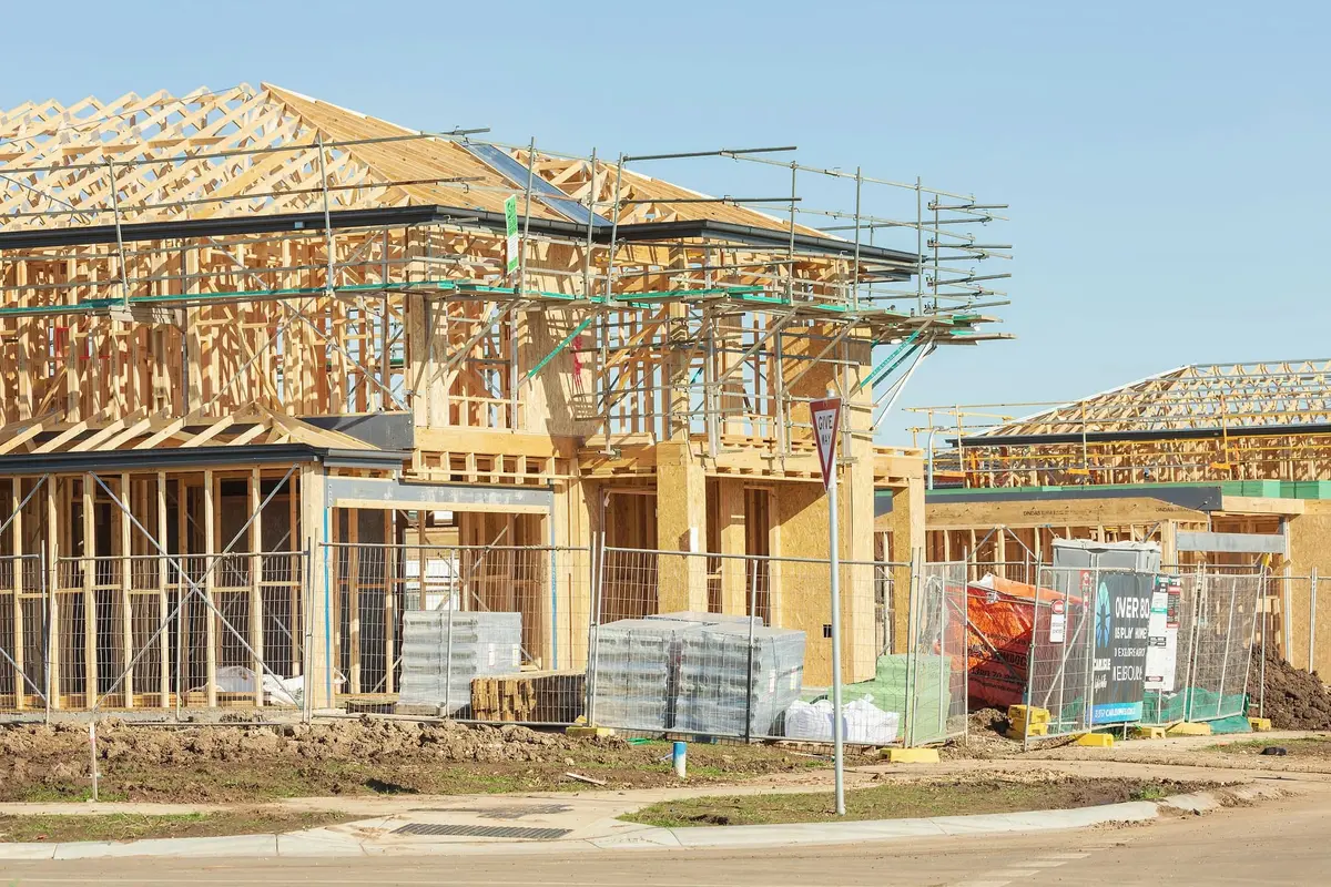 A two-storey timber frame with a complete roof structure and scaffolding on the upper level, seen from the street, with pallets of bricks in front.