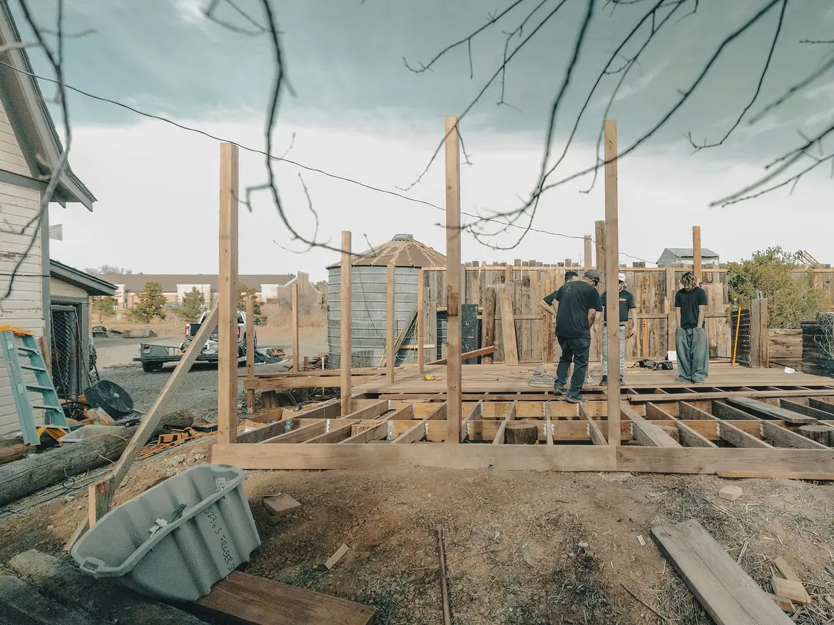 Three builders working on a timber floor deck and the first posts of a wall frame on bare ground, with a grey sky and bare branches above.