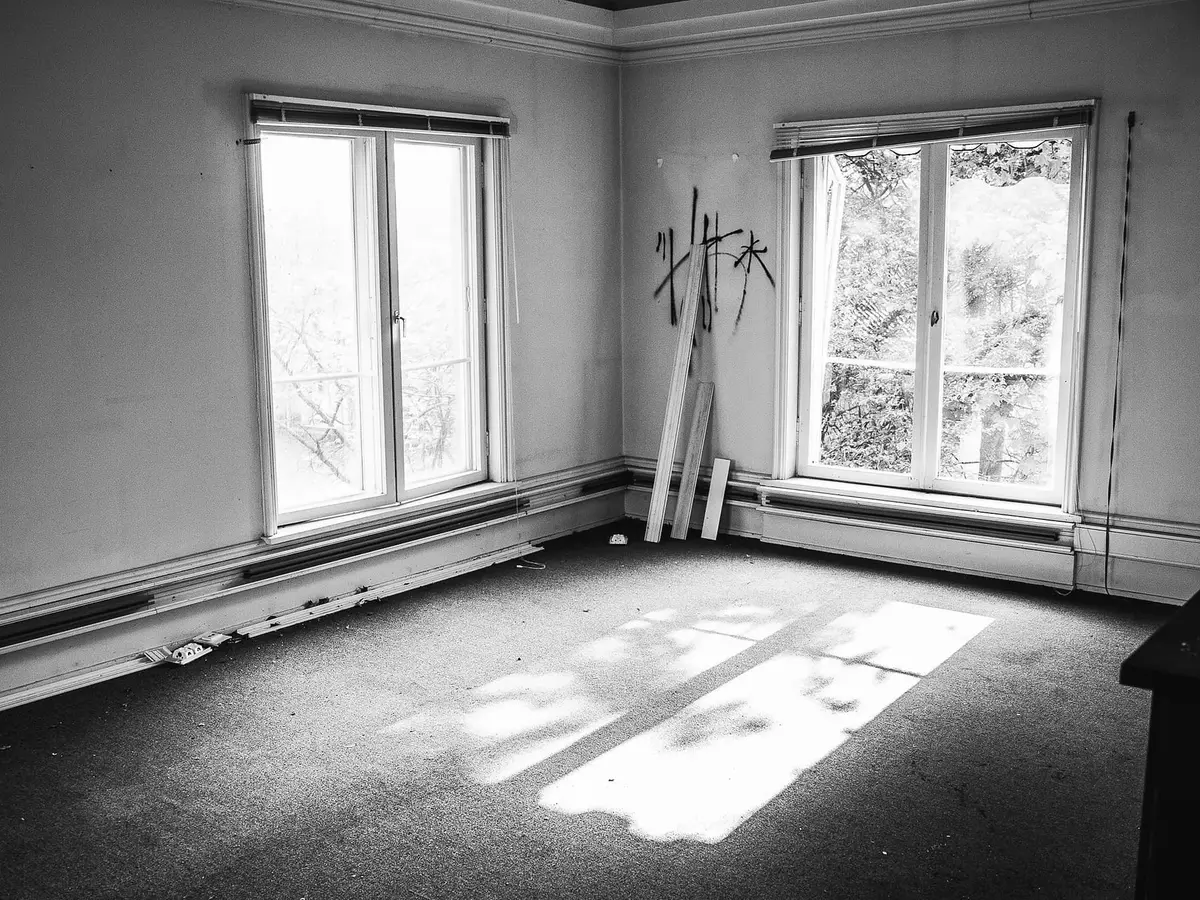 An empty room in black and white with two tall windows, a radiator below them, a stack of timber leaning in the corner and a patch of light on the floor.