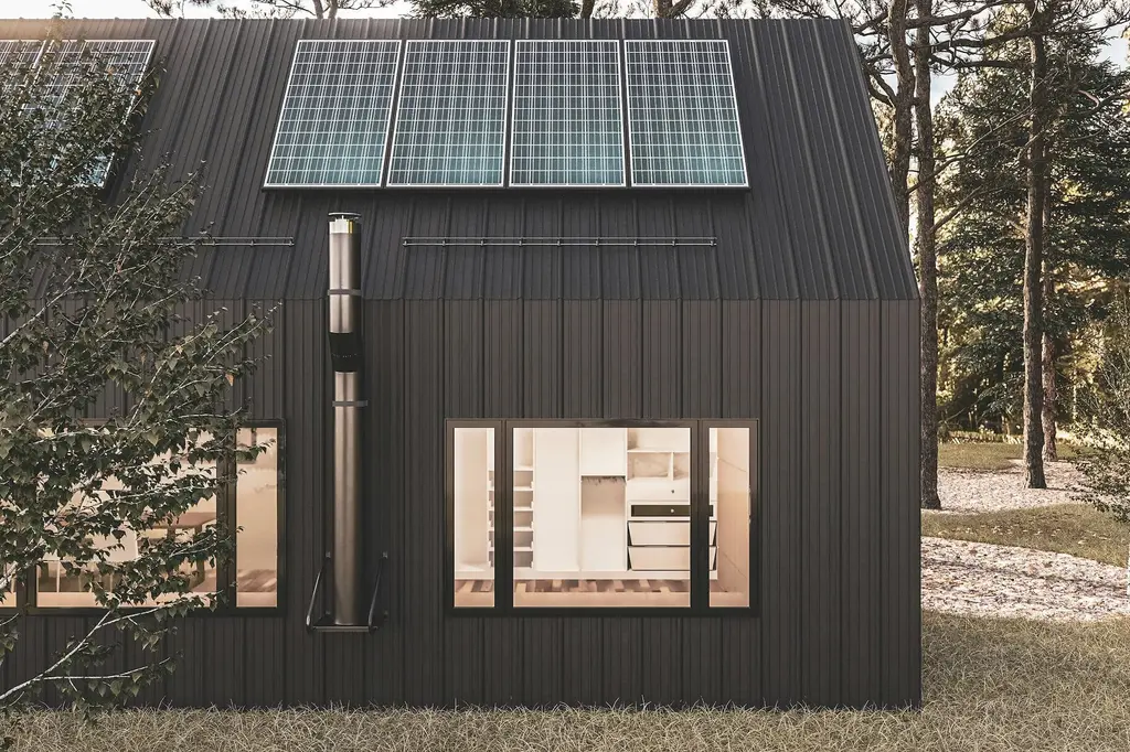 The gable end of a black timber pavilion with solar panels on its roof, a steel flue and a lit window, standing among tall pines.