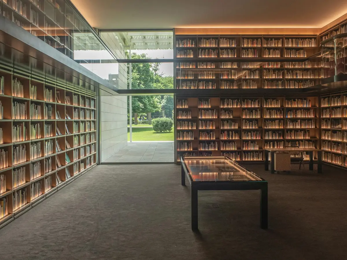 A reading room lined with timber shelves full of books, a long low table in the middle and a glass wall looking onto a lawn.