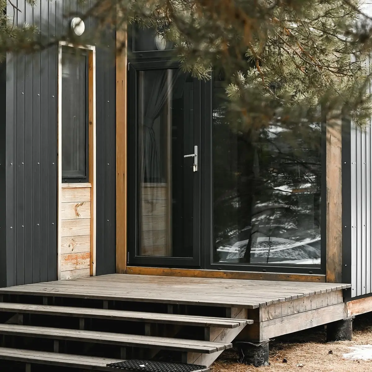 A black boarded wall with a tall door and window, an oak frame and a low timber deck with three steps, under a pine branch.