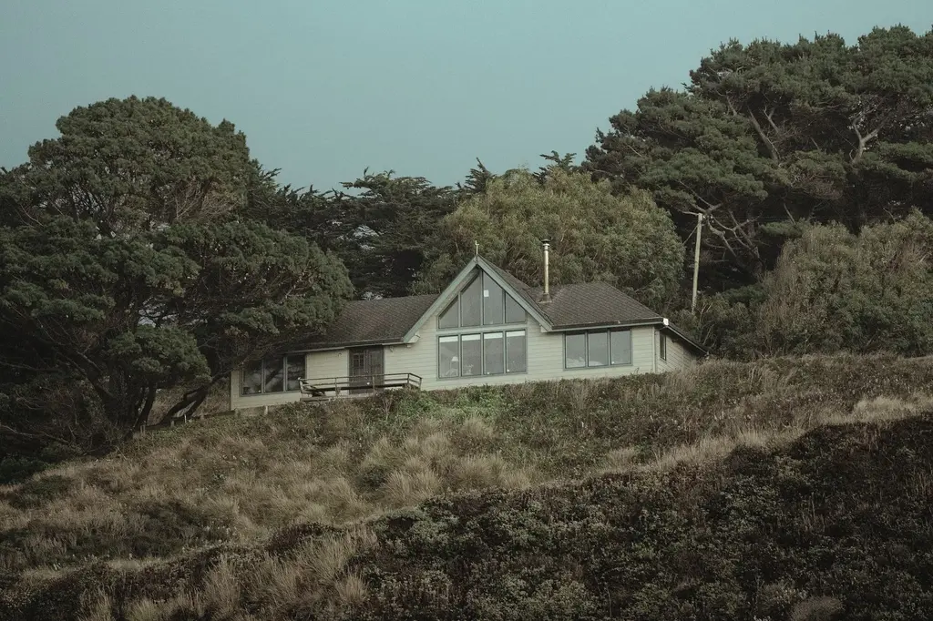 A pale pitched-roof house with a large triangular glazed gable, set on a grassy rise among dark cypress trees.