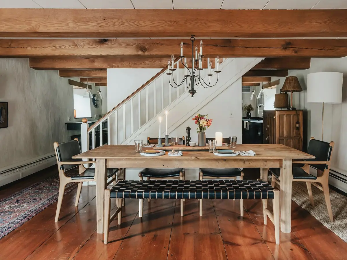 A dining room with exposed oak beams, a white stair behind, a long oak table with a leather bench and a polished wooden floor.