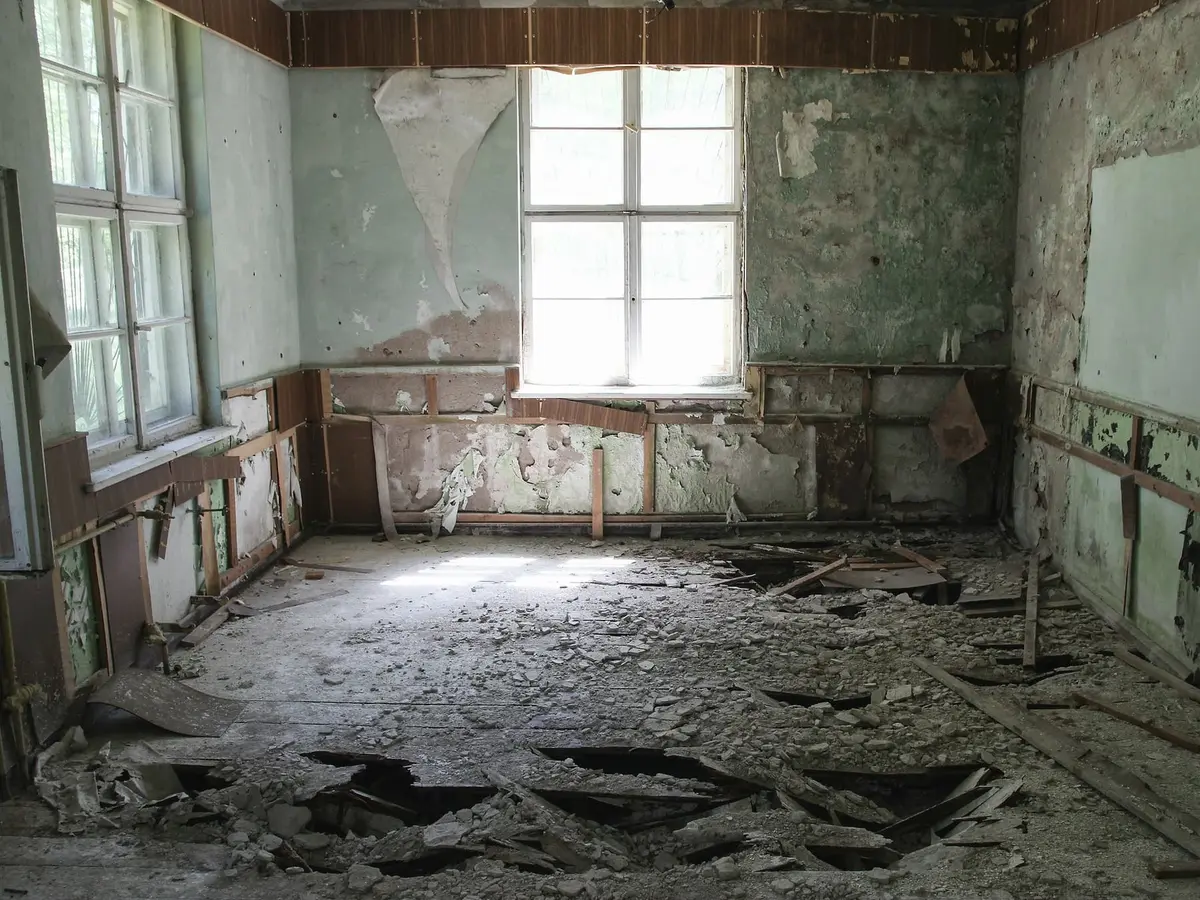 A ruined room with peeling green plaster, a tall window, a broken timber floor and fallen debris.