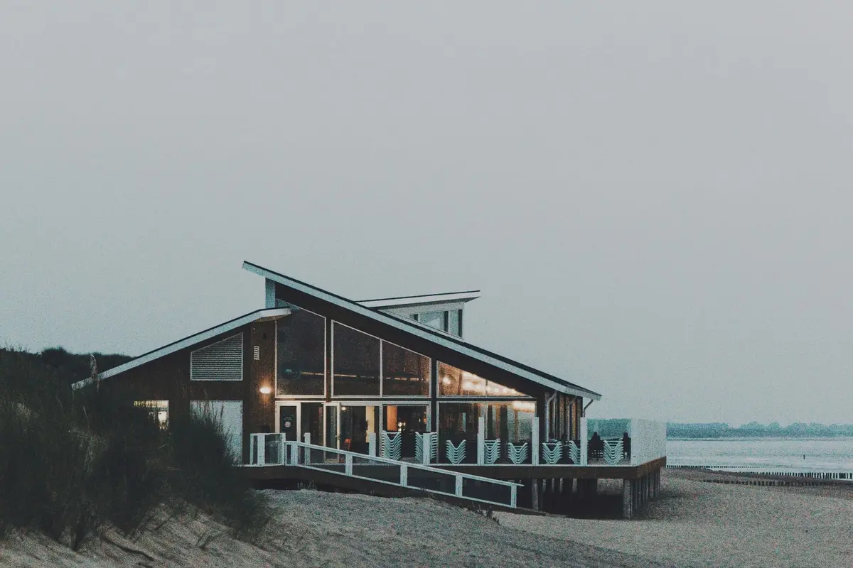 A dark timber beach restaurant with a single steep roof sloping toward the sea, glazed on its long side and lit from inside at dusk.
