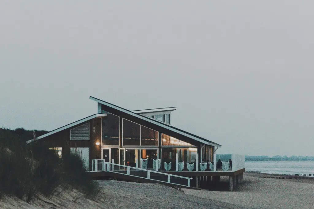 A dark timber beach restaurant with a single steep roof sloping toward the sea, glazed on its long side and lit from inside at dusk.