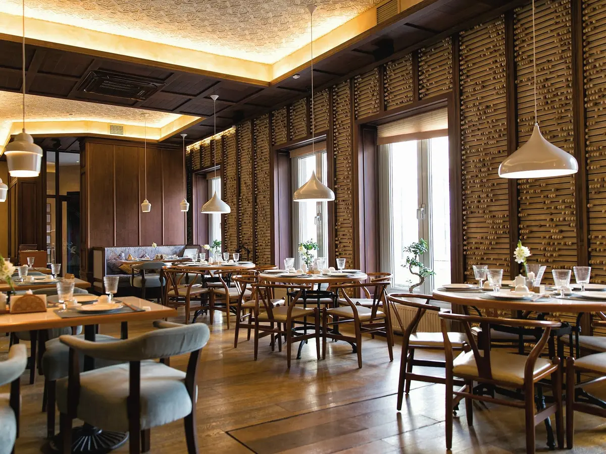 A dining room with timber tables and wishbone-style chairs, round pendant lamps, a warm wood floor and tall windows along one wall.