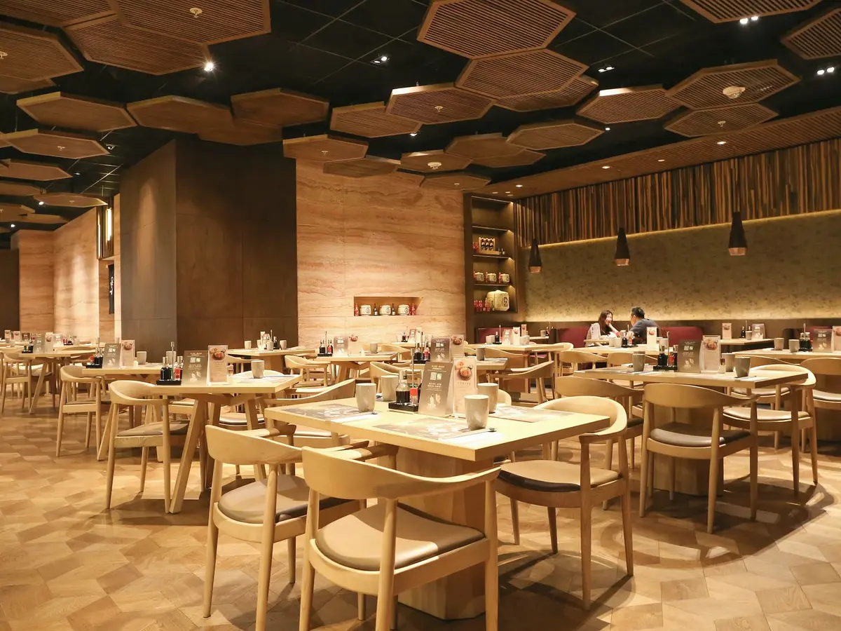 A large restaurant hall with a ceiling of hexagonal timber panels, light wooden tables and chairs and a parquet floor.