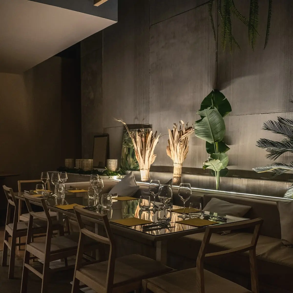 A long dining table laid with glasses and plates in a dim room with a concrete wall, dried grasses in jars and large green leaves.