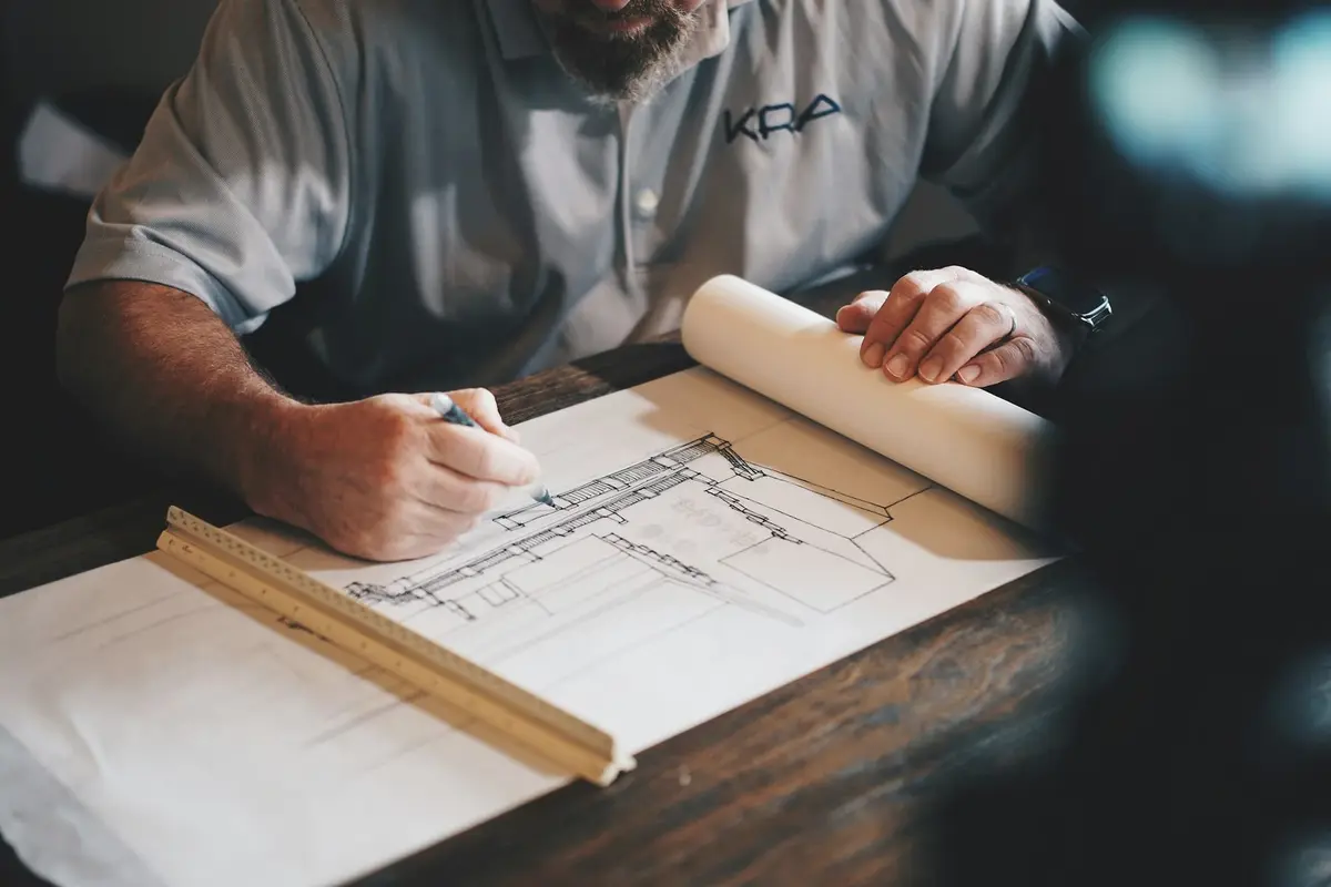 Hands drawing a building section in pencil on tracing paper, with a timber scale rule and a roll of paper beside them.