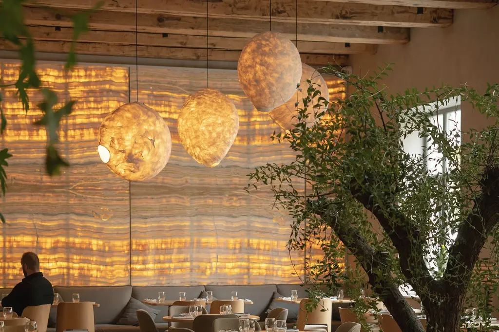 A restaurant with a long wall of glowing amber stone, large round pendant lamps of pale stone, an olive tree in the foreground and low grey banquettes.