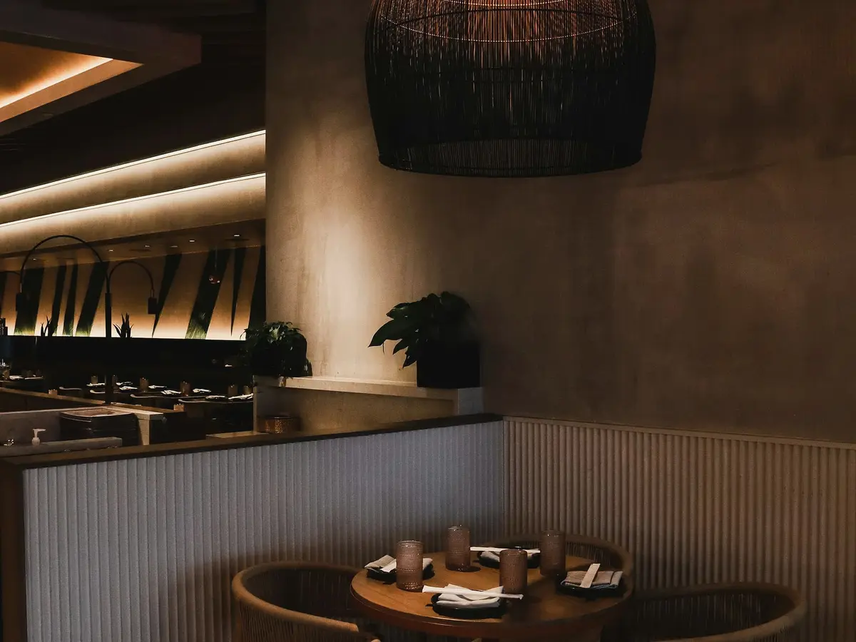 A dim corner of a restaurant with a round table, a fluted pale wall panel, a curved concrete column and a large woven lamp overhead.