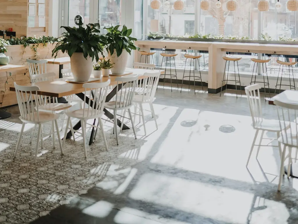 A sunlit room with a long pale table, white spindle-back chairs, patterned floor tiles and potted plants on a window bar.